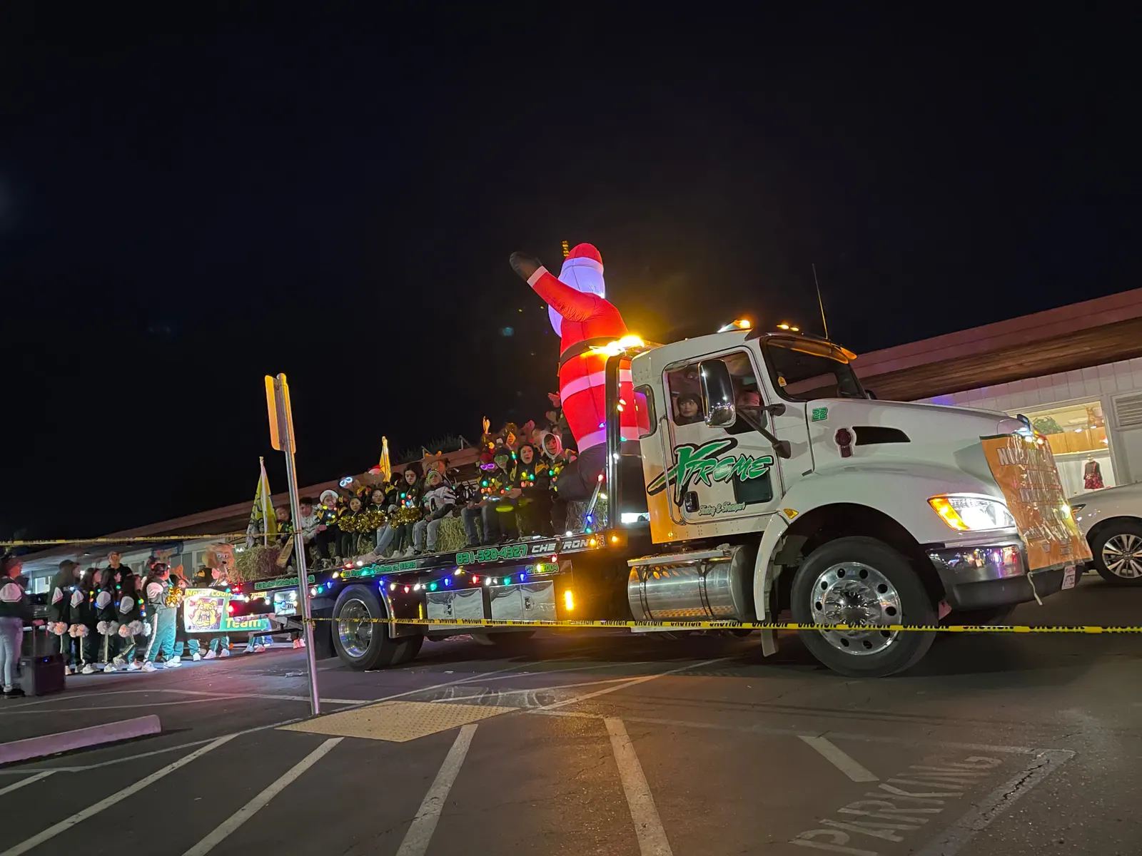 White Xtreme Towing & Transport flatbed truck at night, decorated with holiday lights and an inflatable Santa, carrying a group of young people in a parade.