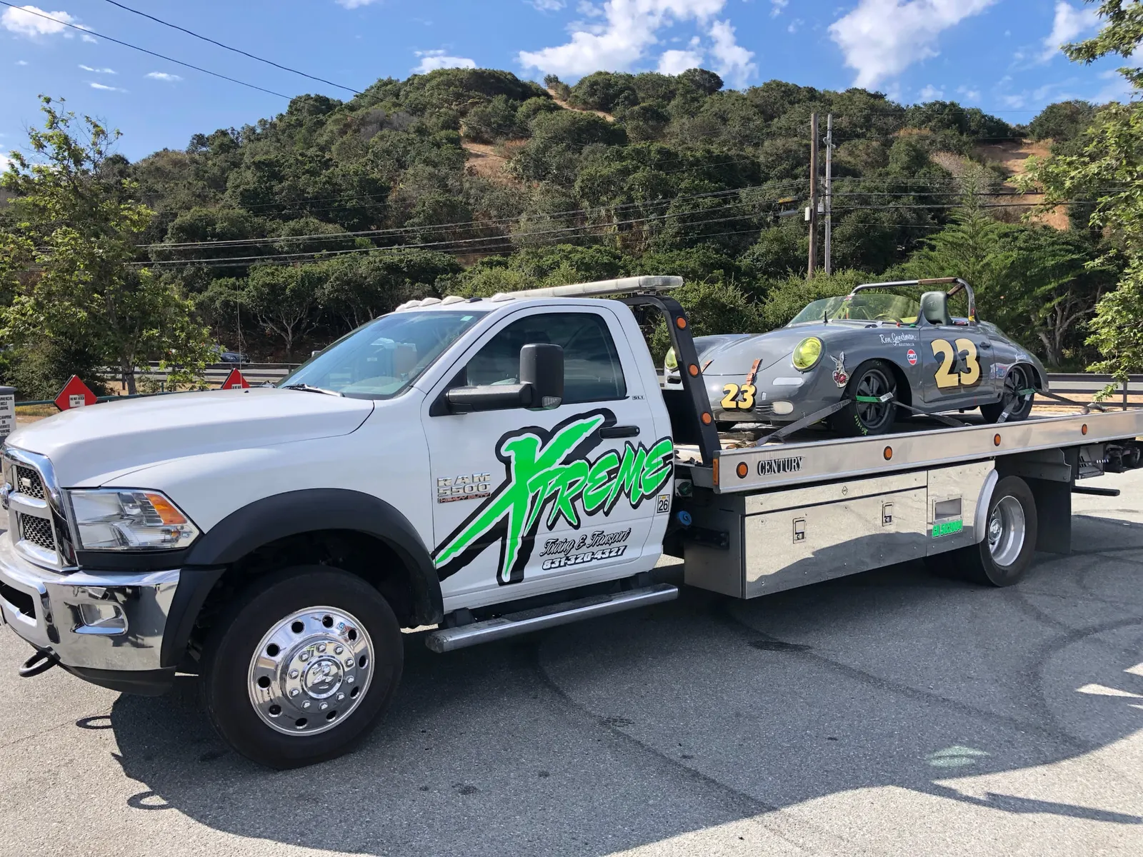 White Xtreme Towing & Transport flatbed truck with a gray number 23 vintage race car strapped on the bed, green hills behind.