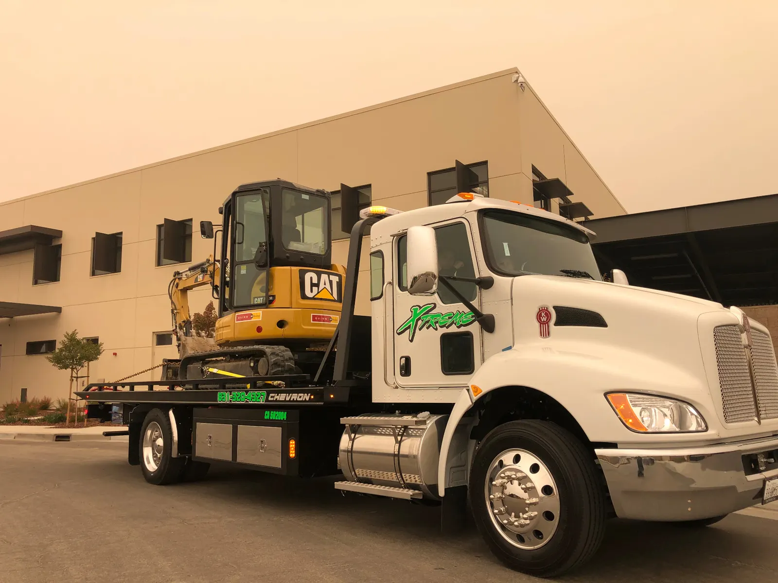 White Xtreme Towing & Transport flatbed truck carrying a yellow compact excavator in front of a beige building.